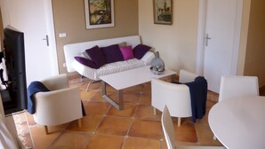 Un salon avec un canapé blanc, deux fauteuils, une table basse et un téléviseur. Il y a deux tableaux au mur et une porte sur la droite.