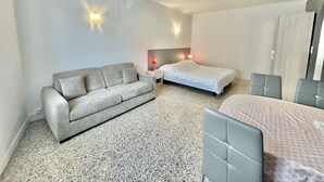 A hotel room with a grey sofa, a single bed, a small bedside table with a lamp, and a wardrobe. - Themis (Cannes)