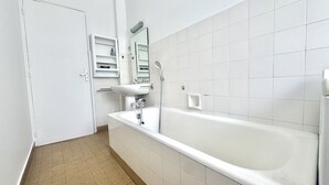 A bathroom with a white bathtub, a sink, and a mirror. There is a door leading to another room and a shelf with toiletries. - Themis (Cannes)