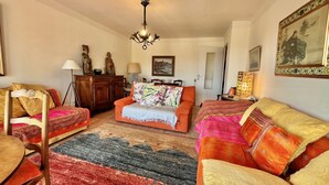 Un salon comprenant un canapé, un fauteuil et une petite table avec une lampe. Un lustre est suspendu au plafond.