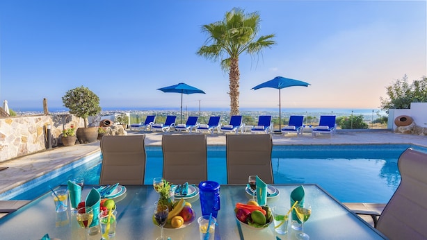 Ein Esstisch gedeckt mit Tellern, Besteck und Getränken mit Blick auf einen Pool mit Liegestühlen und Sonnenschirmen.