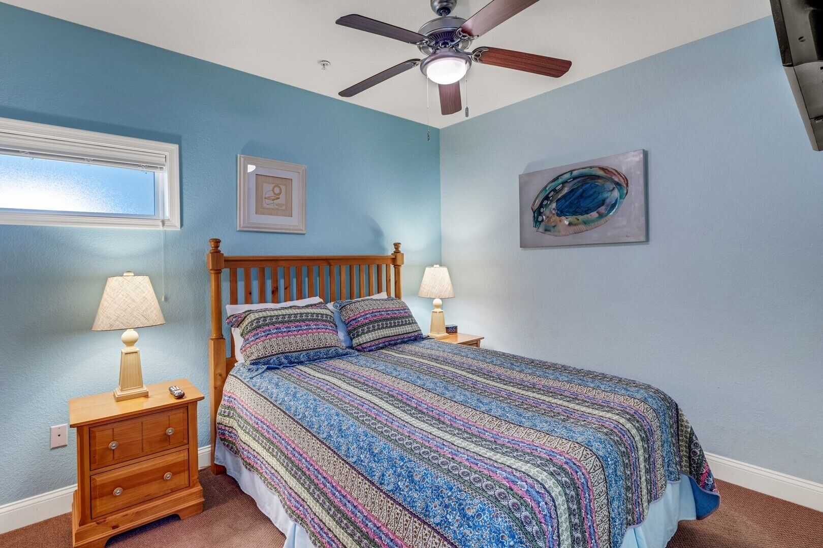 A bedroom with a wooden bed, two bedside tables with lamps, a ceiling fan, a window, and a framed picture on the wall.