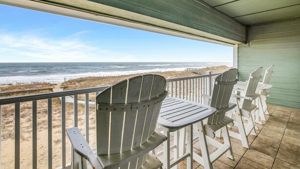 A covered outdoor seating area with Adirondack chairs and a table, overlooking a beach with waves and sand. - FFR211 Atlantic View Retreat by First Flight Rentals (Kill Devil Hills)