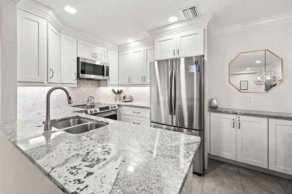 A modern kitchen with white cabinetry, stainless steel appliances, a granite countertop, and a decorative hexagonal mirror. - Tropical Garden View 2 bed, 2 bath at Sanibel Moorings #822 (Sanibel)