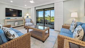 A spacious living room with wicker furniture, a flat-screen TV, and a view of a golf course through large windows. - Newly Renovated Beachfront 3 bed, 2 bath at Sanibel Moorings #711 (Sanibel)