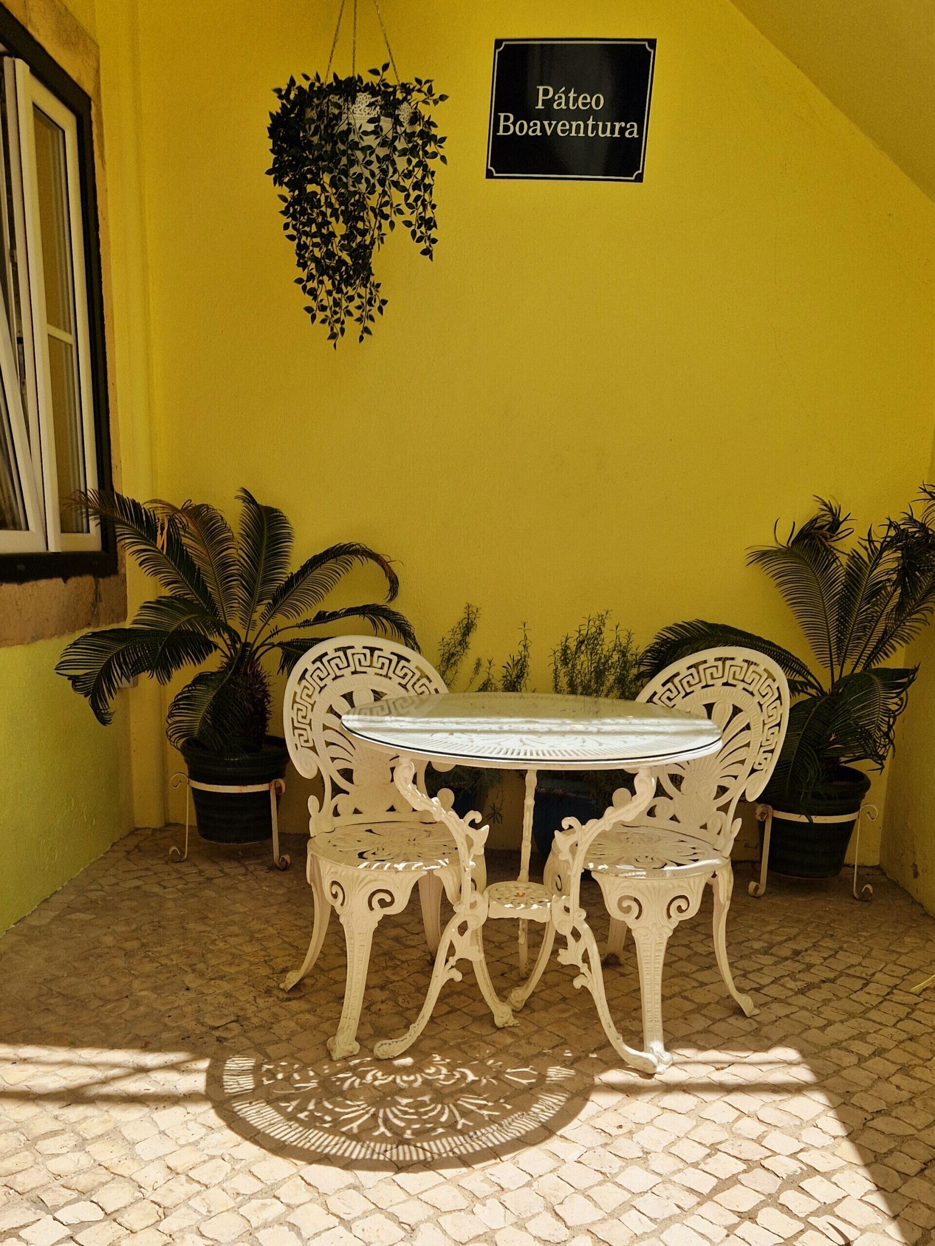 A yellow wall with a hanging plant and a sign that reads "Páteo Boaventura". There is a white table with two chairs in front of it.