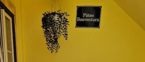 A yellow wall with a hanging plant and a sign that reads "Páteo Boaventura". There is a white table with two chairs in front of it.