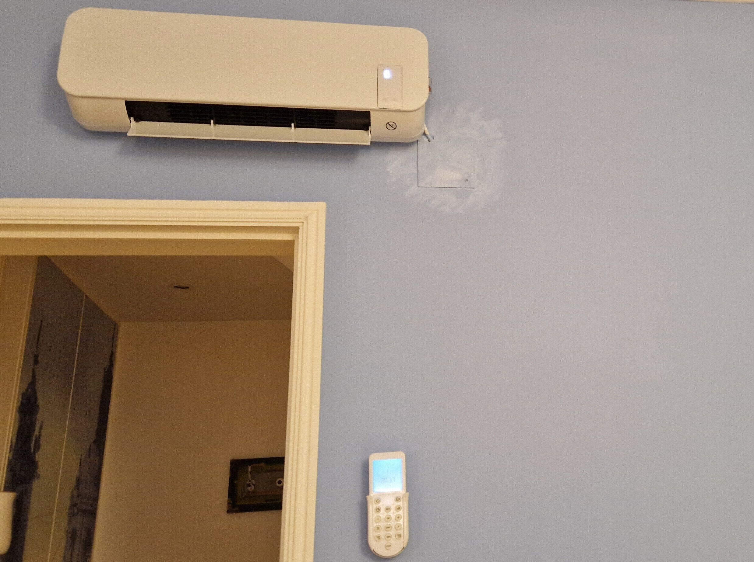 Un climatiseur de couleur beige est fixé sur un mur bleu clair, une télécommande est accrochée au mur, et une porte donne accès à une pièce.