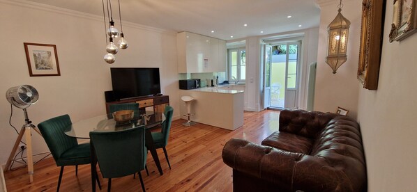 A modern living room with a brown leather sofa, a dining table with chairs, a TV, and a kitchen area visible through an open door.