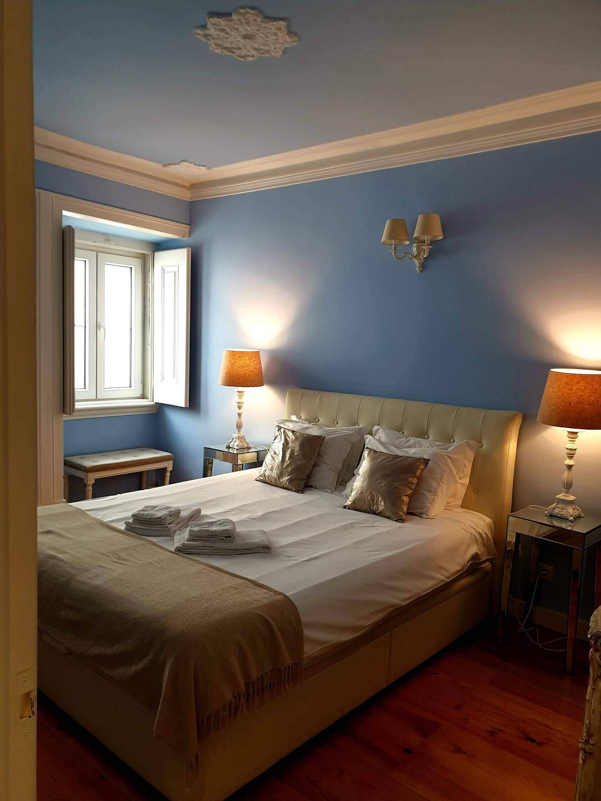 A neatly made bed with white linens and a beige throw blanket, two bedside lamps, a wooden bench, and a window with blinds.