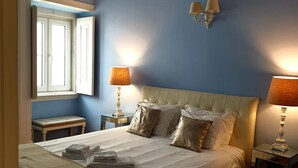 A neatly made bed with white linens and a beige throw blanket, two bedside lamps, a wooden bench, and a window with blinds. - Pateo Boaventura in Bairro Alto (Lisbon)