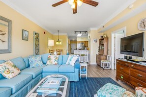 A living room with a sofa, coffee table, TV, and a kitchen in the background. - SP202S - Sea Grace by First Flight Rentals (Kill Devil Hills)