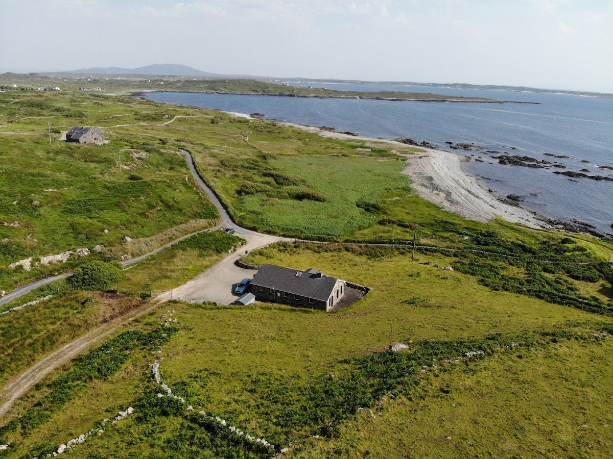 Vue aérienne d’une propriété située sur la côte, comprenant un bâtiment principal, une dépendance et une route menant à la plage.