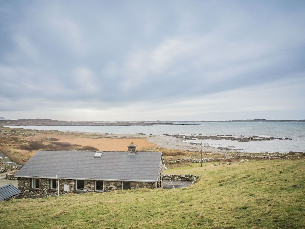 Une maison en pierre avec un toit en pente, située sur une colline verdoyante qui offre une vue sur une plage et la mer.