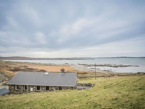 A stone house with a sloped roof, situated on a grassy hill overlooking a beach and the sea.