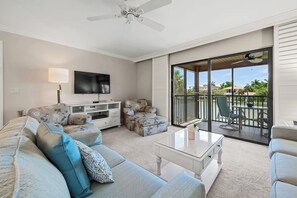 Ein geräumiges Wohnzimmer mit Flachbildfernseher, Deckenventilator, Glastisch und einem Balkon mit Gartenmöbeln.