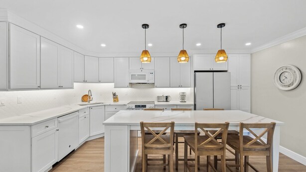 Une cuisine moderne avec des meubles blancs, un îlot central doté de chaises en bois et des luminaires suspendus.