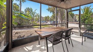 Une terrasse couverte avec une table en bois et des chaises en osier noir, de grandes fenêtres offrant une vue sur les palmiers et les voitures garées.