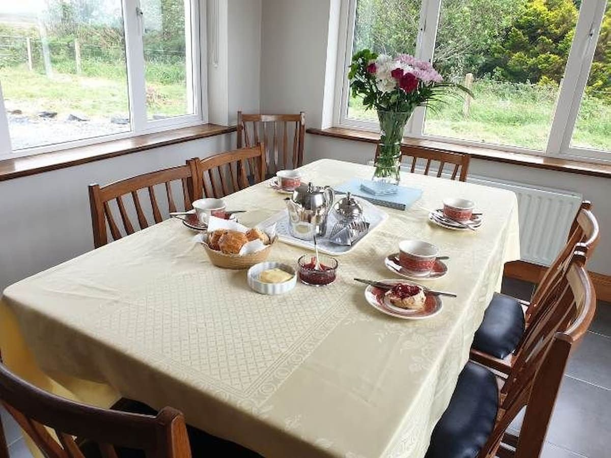Mesa lista para una comida, con sillas alrededor, un florero con flores y una ventana con vista a follaje verde.