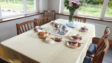 Mesa lista para una comida, con sillas alrededor, un florero con flores y una ventana con vista a follaje verde.