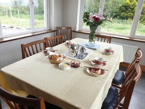 A dining table set for a meal with chairs around it, a vase with flowers, and a window with a view of greenery. - Hillside House (Clifden)