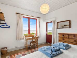A bedroom with a bed, a desk, a chair, a dresser, and a view of the outdoors. - Salmon Cottage (Cashel)
