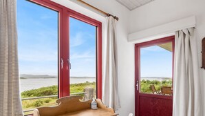 Een kamer met een houten tafel en stoelen, een rode deur die leidt naar een buitenruimte met uitzicht op de zee, en een raam met gordijnen.