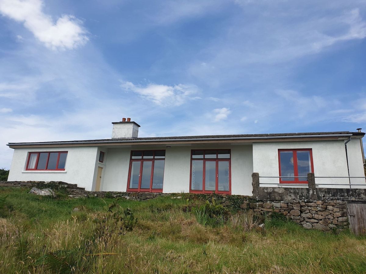 Einbungalow mit weißer Fassade und roten Fensterrahmen, gelegen auf einem grasbewachsen Hügel unter blauem Himmel.