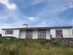 A single-story house with a white exterior and red-framed windows, situated on a grassy hill with a clear sky.