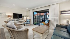A living room with a television, a sofa, a coffee table, and a chair. There is a balcony with a view of buildings. - Gorgeous Partial Beach View 3 bed, 2 bath at Sanibel Moorings #532 (Sanibel)