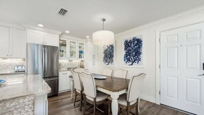 A modern kitchen with white cabinetry, a stainless steel refrigerator, a dining table with chairs, and two wall-mounted artworks. - Gorgeous Partial Beach View 3 bed, 2 bath at Sanibel Moorings #532 (Sanibel)