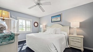 Une chambre à coucher comprenant un lit, deux tables de chevet, un ventilateur de plafond, une fenêtre avec des stores, un miroir fixé au mur et un tableau encadré.