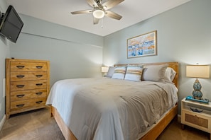 A bedroom with a bed, wooden dresser, ceiling fan, wall-mounted TV, and a painting. - FFR110 - Nautical Twilight by First Flight Rentals (Kill Devil Hills)