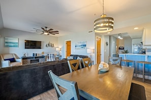 A modern living room with a dark sofa, a wooden dining table, and a kitchen area with a bar. - FFR110 - Nautical Twilight by First Flight Rentals (Kill Devil Hills)