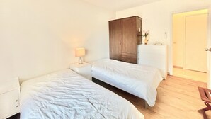 Een slaapkamer met twee eenpersoonsbedden, een houten garderobe, een nachtkastje met een lamp, en een deur naar een andere kamer.