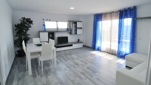 A modern kitchen with white cabinetry, a dining table with chairs, and a large window with blue curtains. - HomeHolidaysRentals Solmar - Costa Barcelona (Santa Susana)