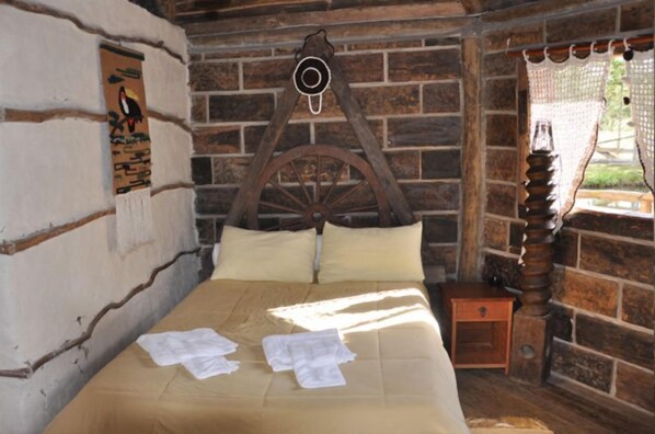 Uma cama de madeira rústica com cabeceira, uma pequena mesa de cabeceira de madeira e um relógio fixado na parede.