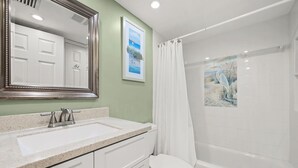 Une salle de bain avec un lavabo, un meuble-lavabo et des toilettes, tous de couleur blanche. On y trouve un rideau de douche et un tableau encadré accroché au mur.