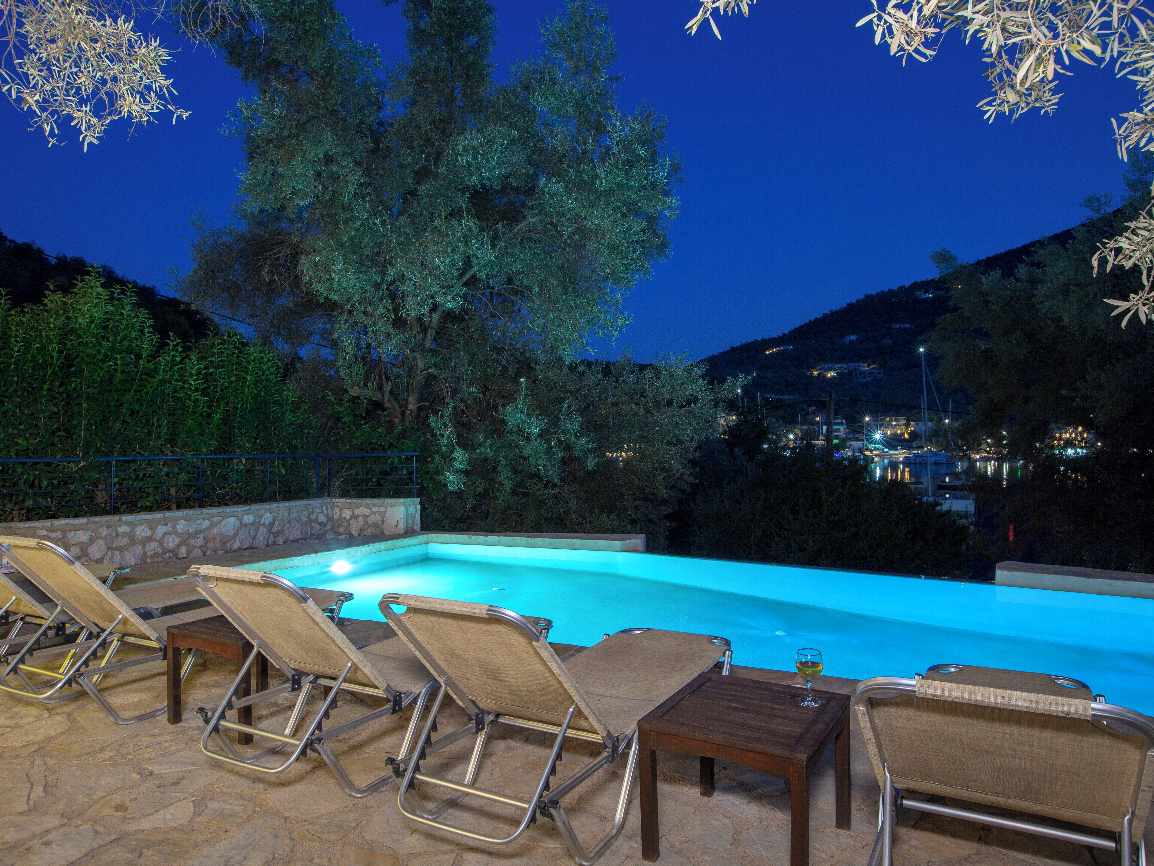 A pool area with lounge chairs and a table, surrounded by trees and a view of a town at night.