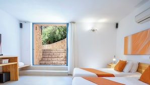 Une chambre d’hôtel moderne avec deux lits, un bureau en bois, une grande fenêtre donnant sur un escalier en pierre et de la verdure, et un climatiseur fixé au mur.
