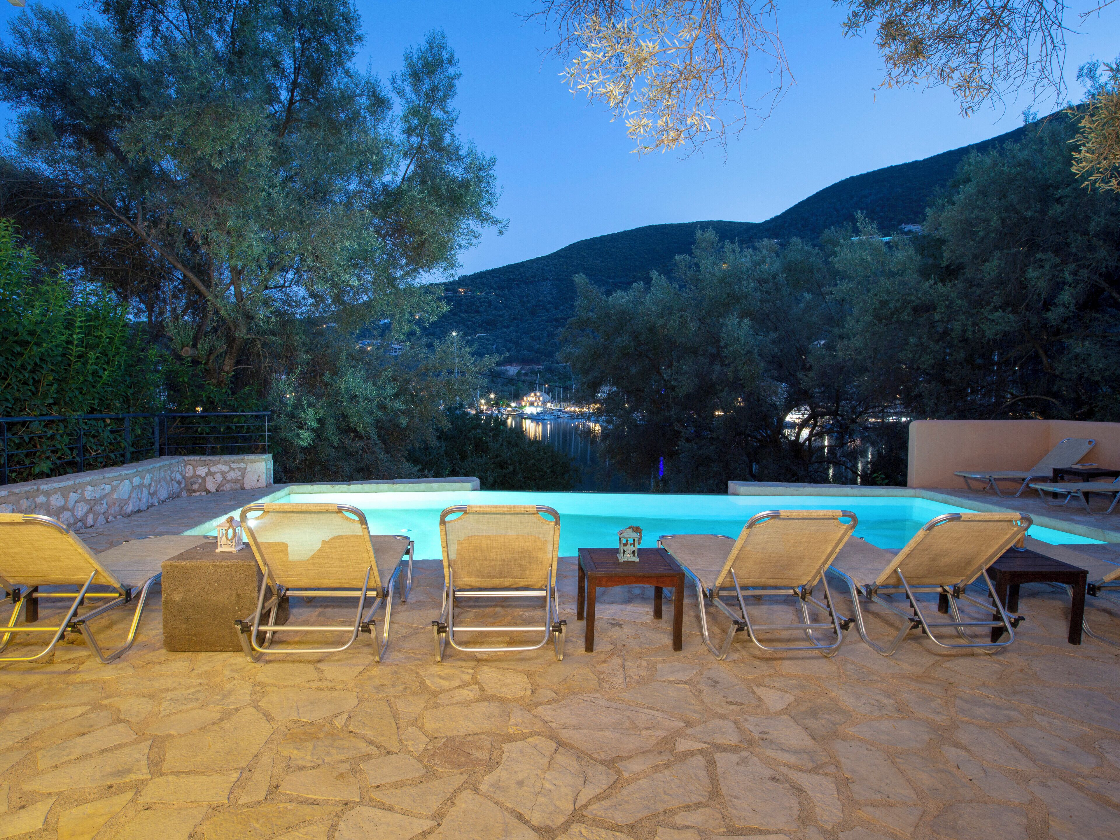 A poolside area with lounge chairs and a stone-tiled floor, overlooking a body of water and hills.