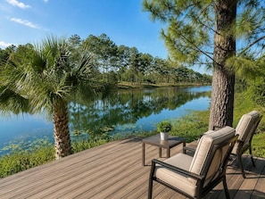 Una terraza de madera con mobiliario exterior, un lago tranquilo y árboles circundantes.