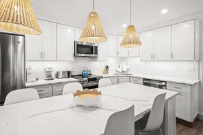 Une cuisine moderne avec des meubles blancs, un réfrigérateur en acier inoxydable, un four à micro-ondes, un grille-pain et une bouilloire. On y trouve trois lampes suspendues en osier, une table à manger blanche avec des chaises, et un bol décoratif posé sur la table.