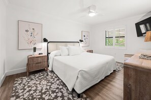 Ein Schlafzimmer mit Bett, Nachttischen, Deckenventilator, Kunstwerken und einem Fenster mit Jalousien.