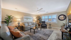 Sala de estar com sofá, poltronas, mesa de centro e luminária de chão. Há um ventilador de teto, um quadro na parede e uma janela com persianas.
