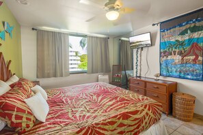 Ein Schlafzimmer mit einem Bett, einem Deckenventilator, einem Fernseher, einem Gemälde und einem Fenster mit Vorhängen.