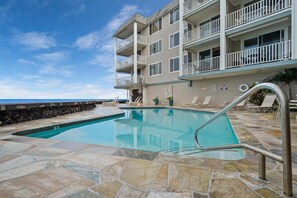 A swimming pool with a stone deck, a building with balconies, and a view of the ocean.