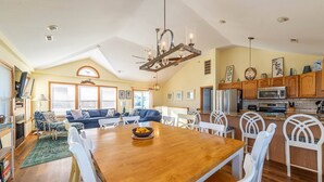 A spacious living room with a wooden dining table, white chairs, and a chandelier.