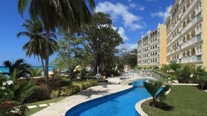 Area piscina di un resort con una piscina blu cristallina, palme e un edificio a più piani con balconi.