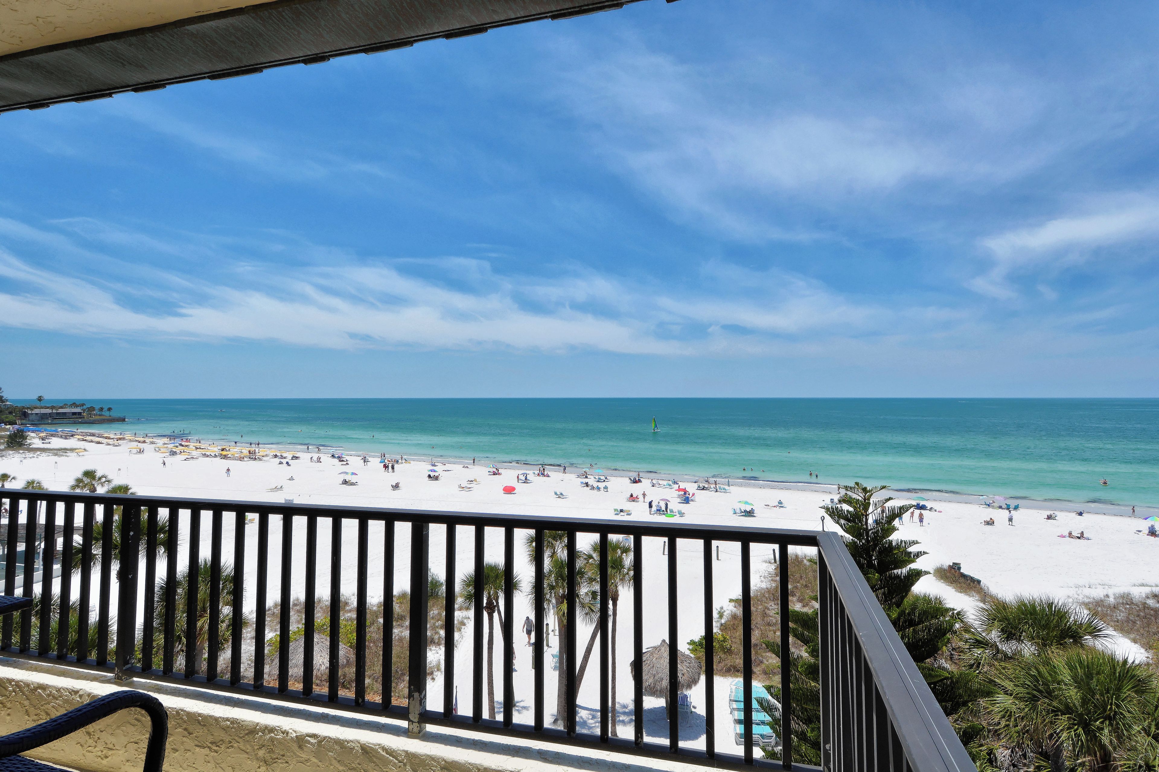 A beach with white sand and clear blue water, viewed from a balcony with a railing.